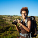beautiful african girl with backpack smiling, holding binoculars, canyon background.