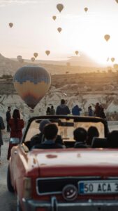 Scenic view of hot air balloons rising at sunrise in Cappadocia with people watching from a convertible car.